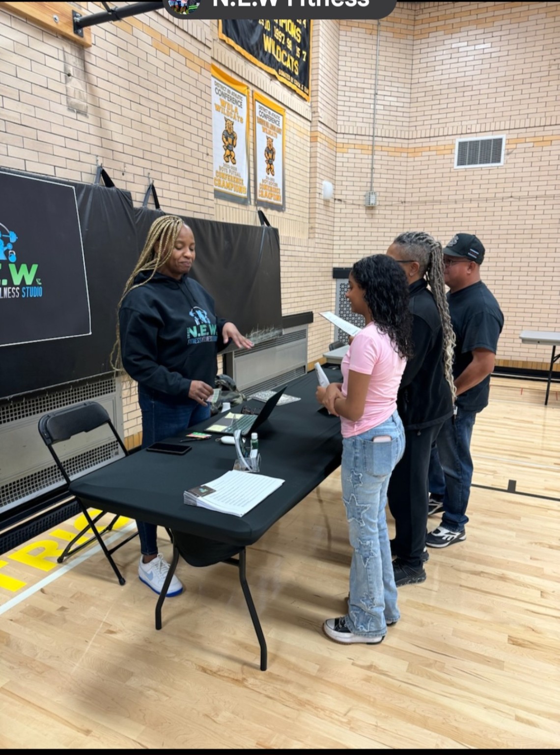 Coach T speaking with young people at the N.E.W. Fitness table in a school gym