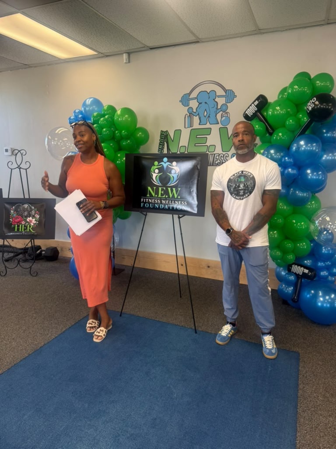 Two people beside the N.E.W. Fitness Wellness Foundation display and blue and green balloons