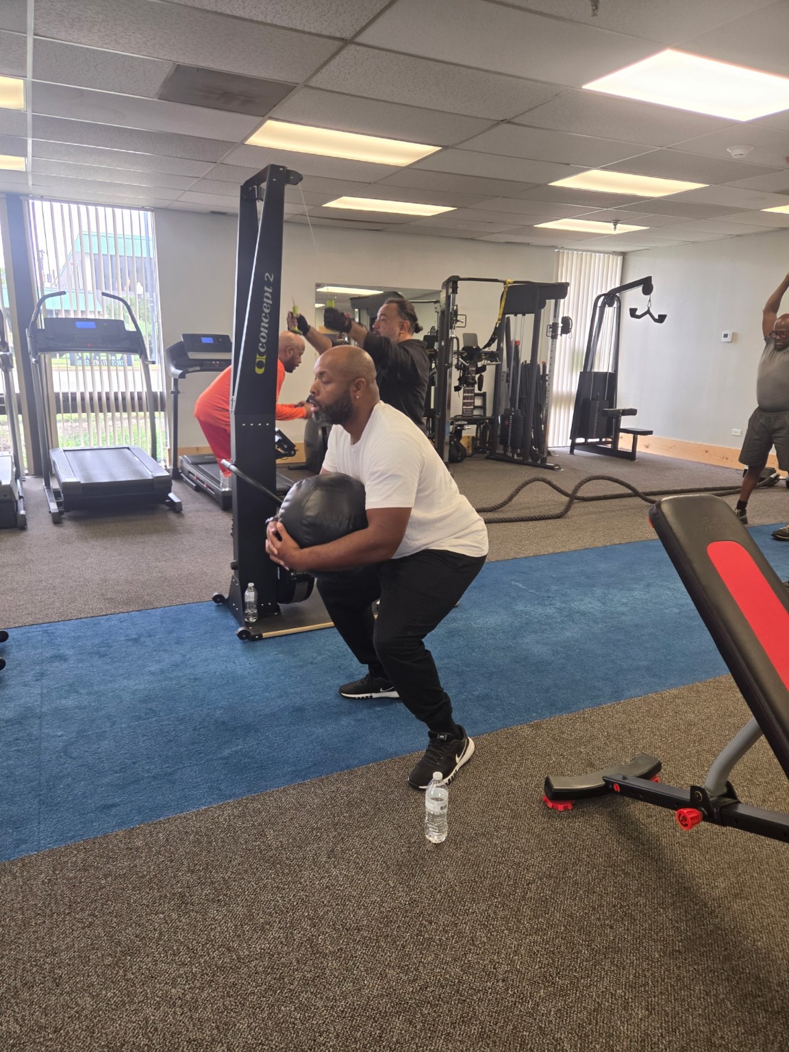 A man holding a medicine ball in a squat while other men exercise in the studio